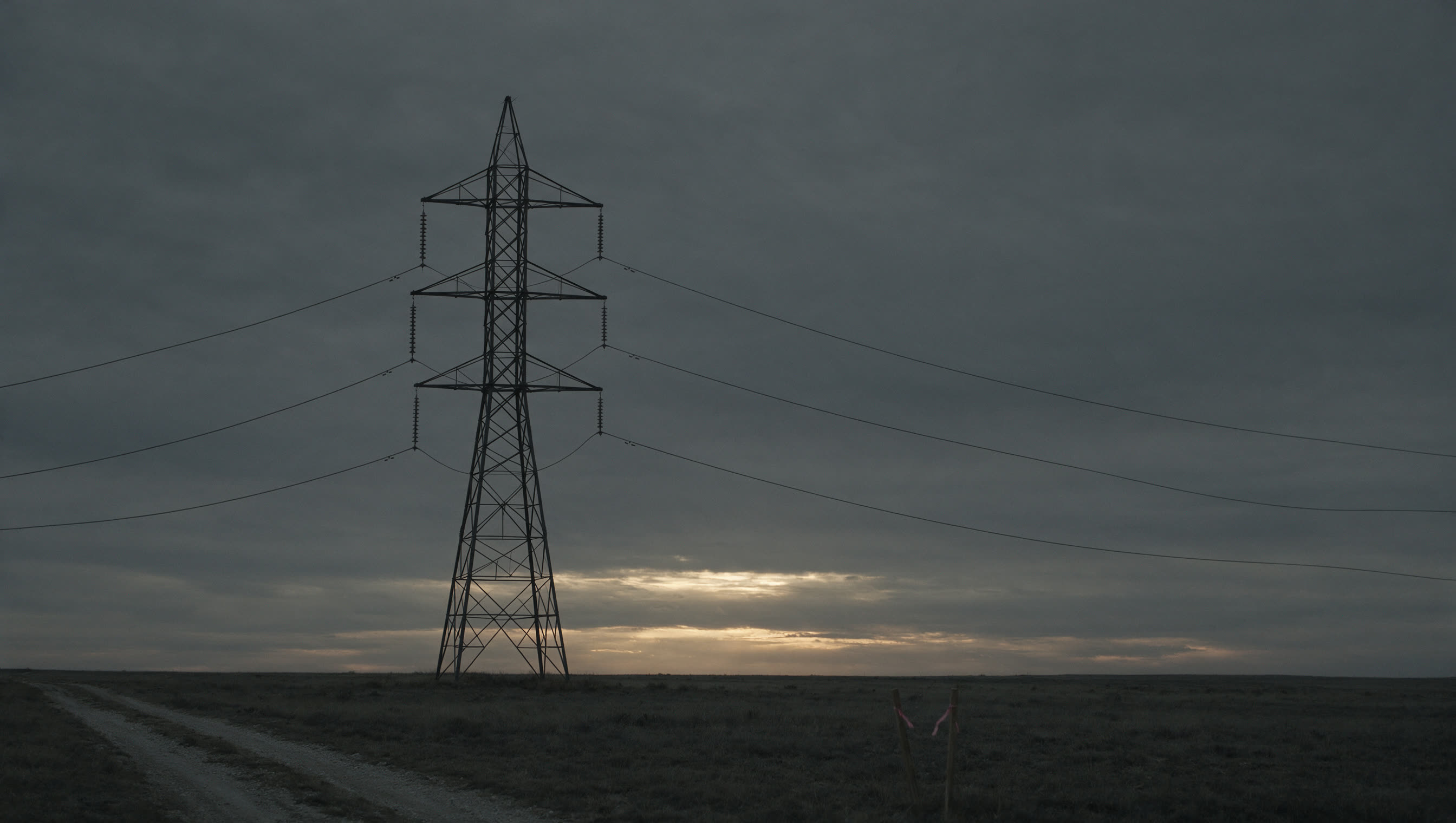 A steel lattice transmission tower standing alone at dusk over open scrub, a caliche two-track running past it and surveyor's stakes in the foreground.