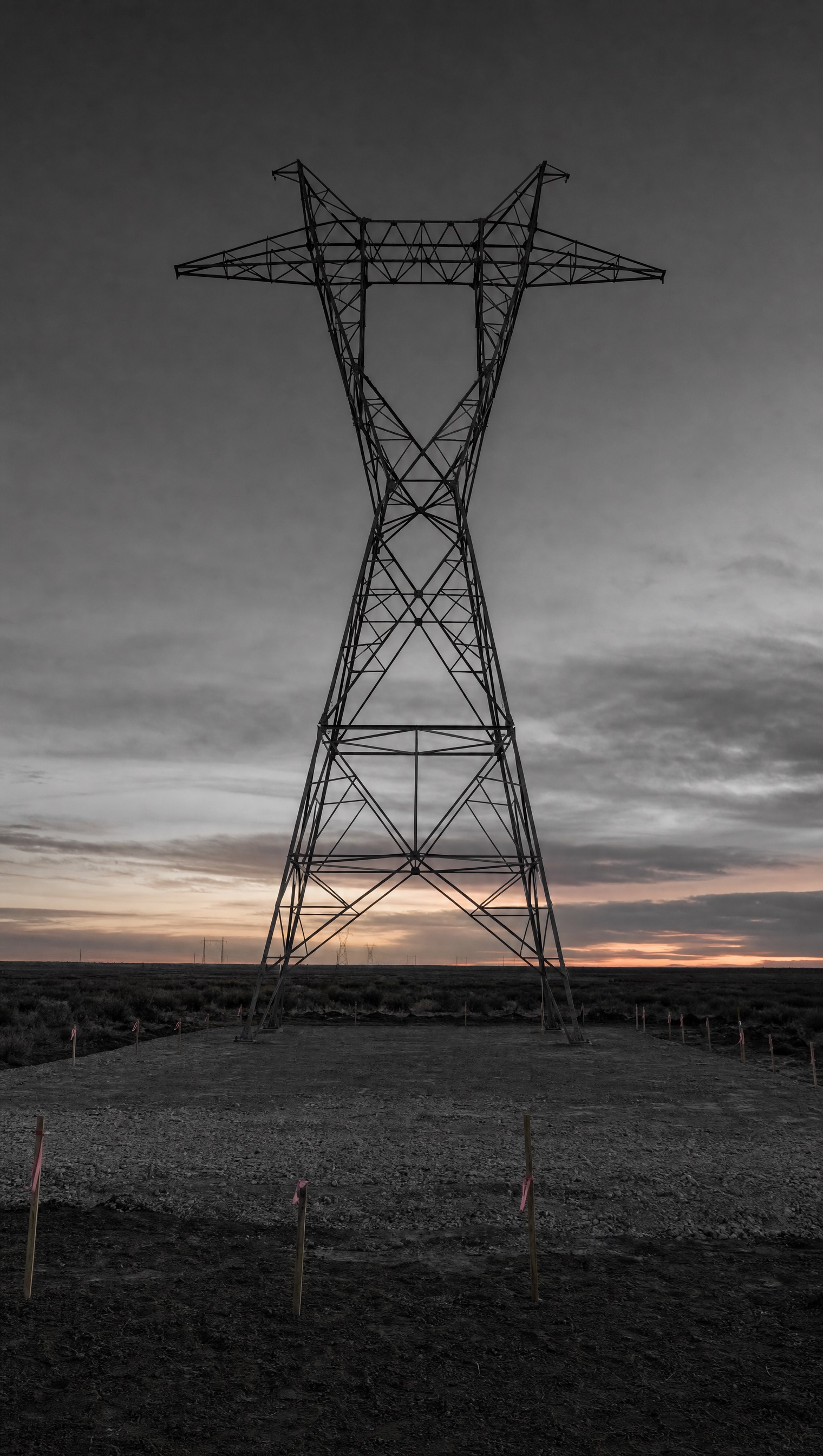 A single lattice transmission tower standing dead centre against a clouded sunset, above a freshly graded pad ringed with surveyor's stakes and pink flagging.